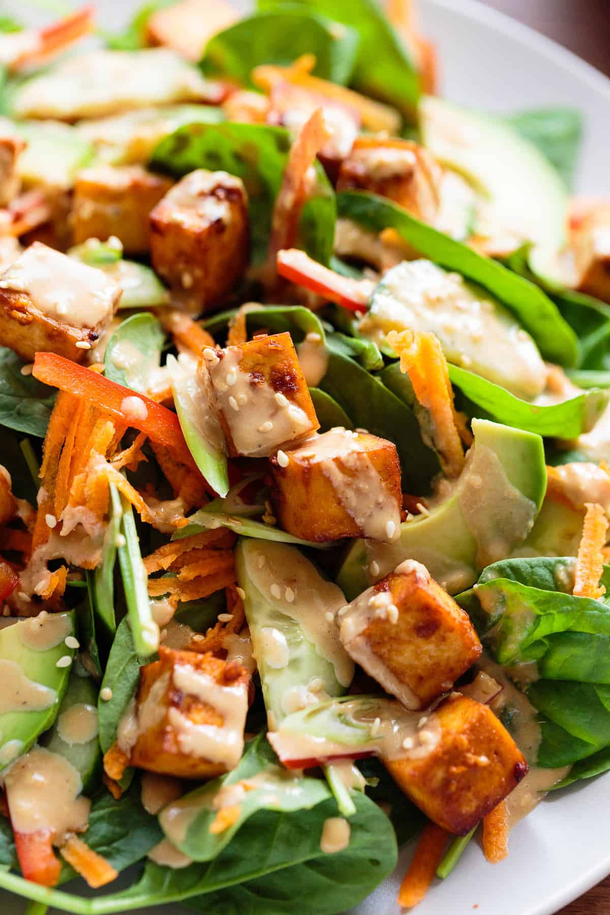 Close up shot of Salad of baby spinach, marinated tofu, avocado, carrot, cucumber, red bell pepper, green onion with a creamy toasted sesame and soy dressing.