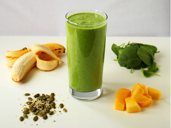 Smoothie in a glass surrounded by key ingredients: spinach, pumpkin seeds, bananas, hemp seeds.