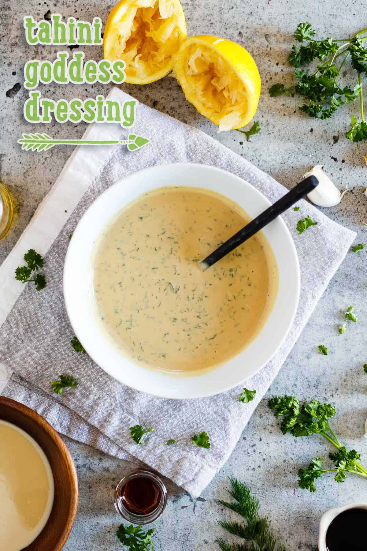 A bowl of Vegan Tahini Goddess Dressing surrounded by key ingredients on a concrete background with typography overlay.