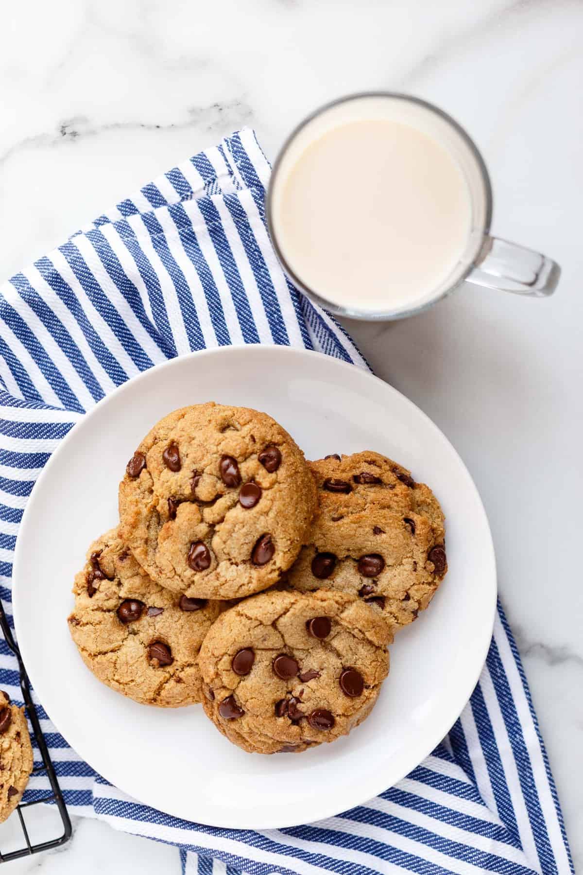 Close up of a plate of vegan chocolate chip cookies on a blue striped tea towel with a glass a non-dairy milk on a marble countertop.