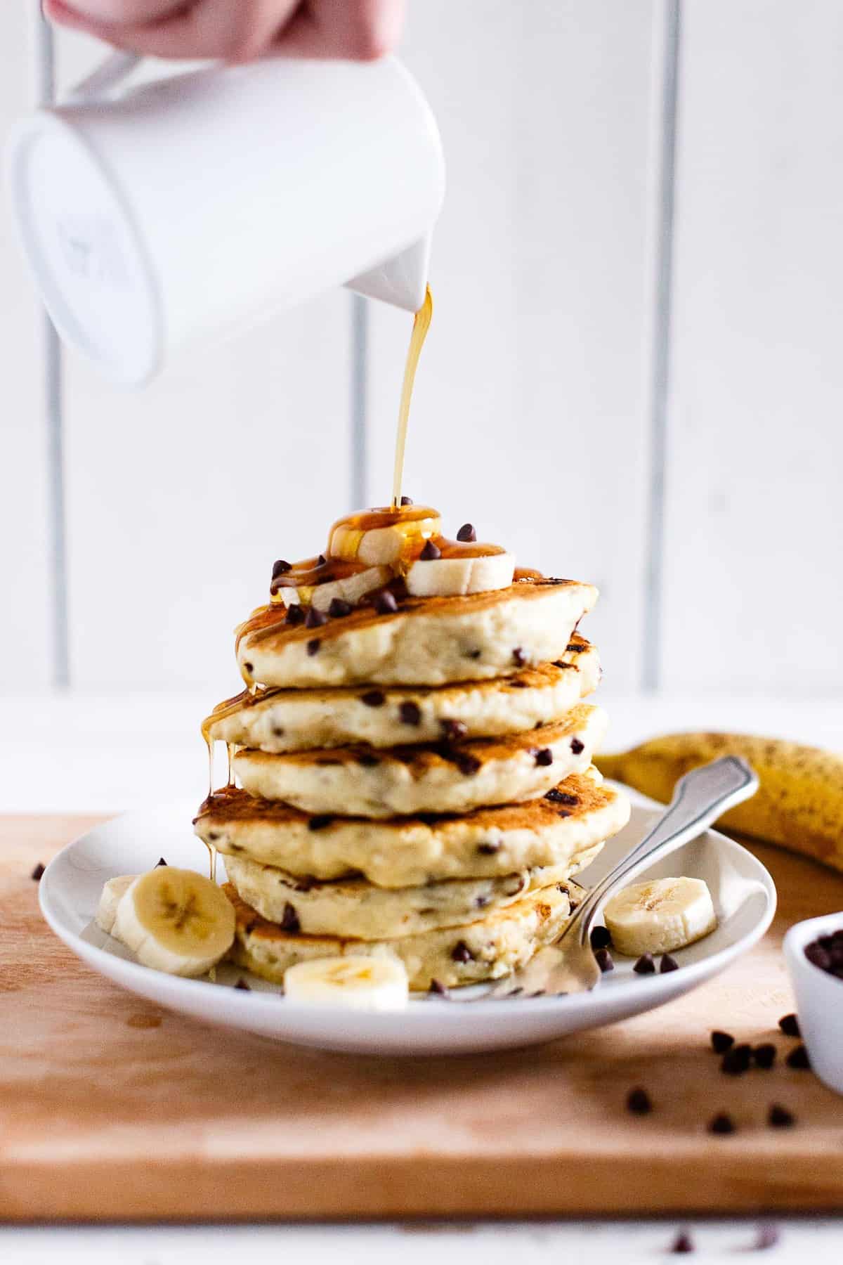 Pouring maple syrup on a stack of vegan chocolate chip pancakes topped with sliced bananas.