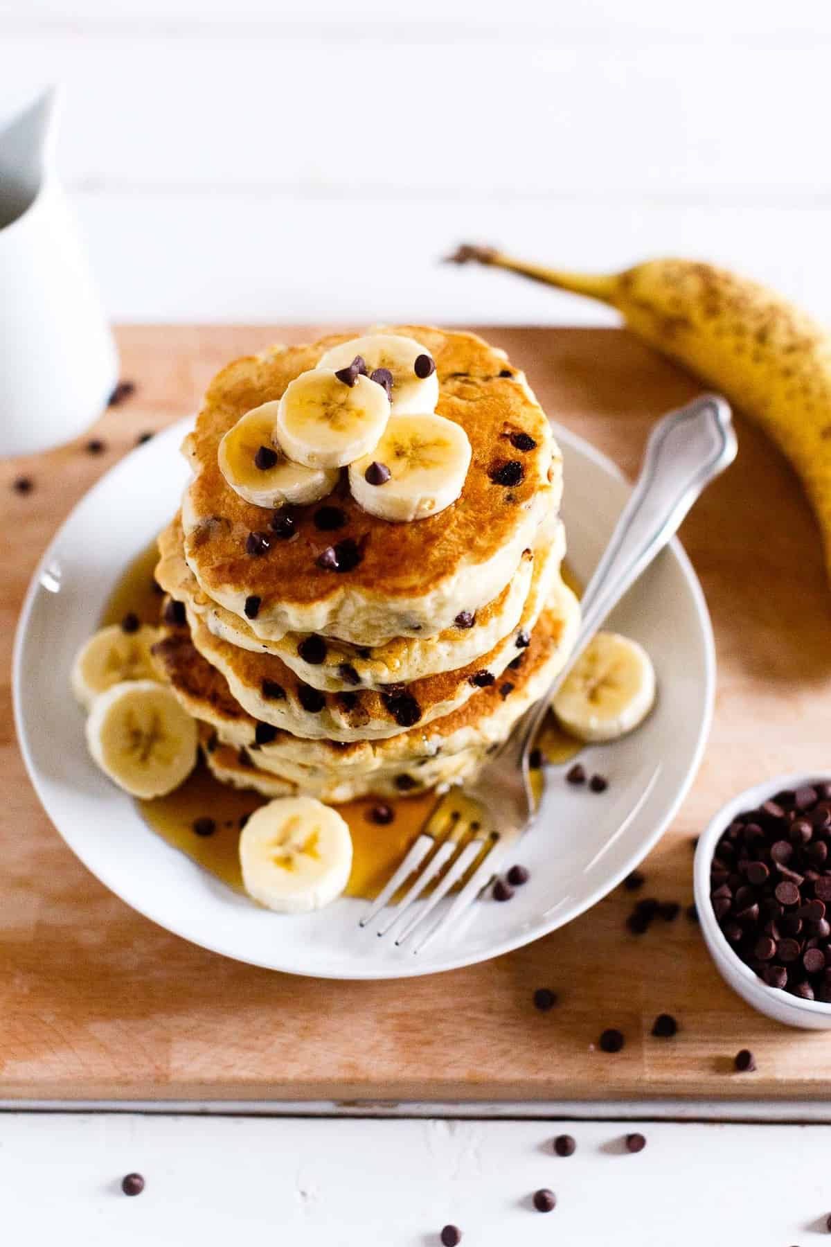 Stack of vegan chocolate chip pancakes topped with sliced bananas and maple syrup.