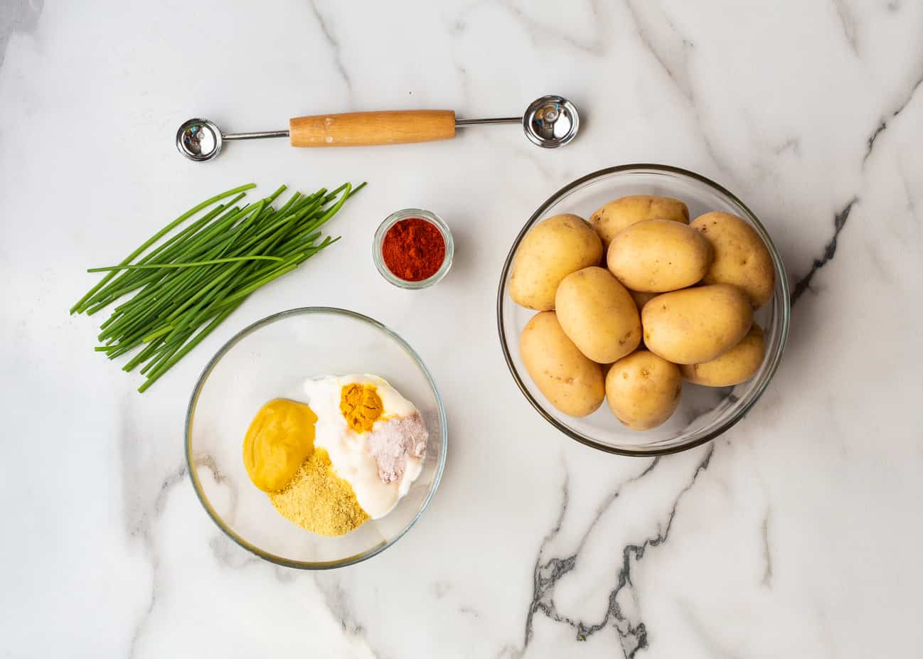 Ingredients for making deviled egg potatoes: Creamer potatoes, fresh chives, paprika, a bowl containing vegan mayo, nutritional yeast, mustard, vinegar, and black salt.