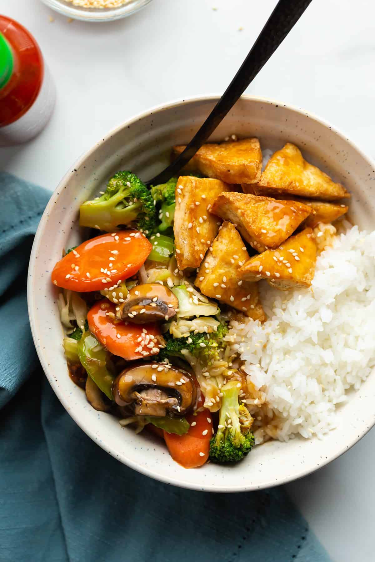 A bowl of steamed rice, tender-crisp vegetables and golden tofu topped with teriyaki sauce and toasted sesame seeds.