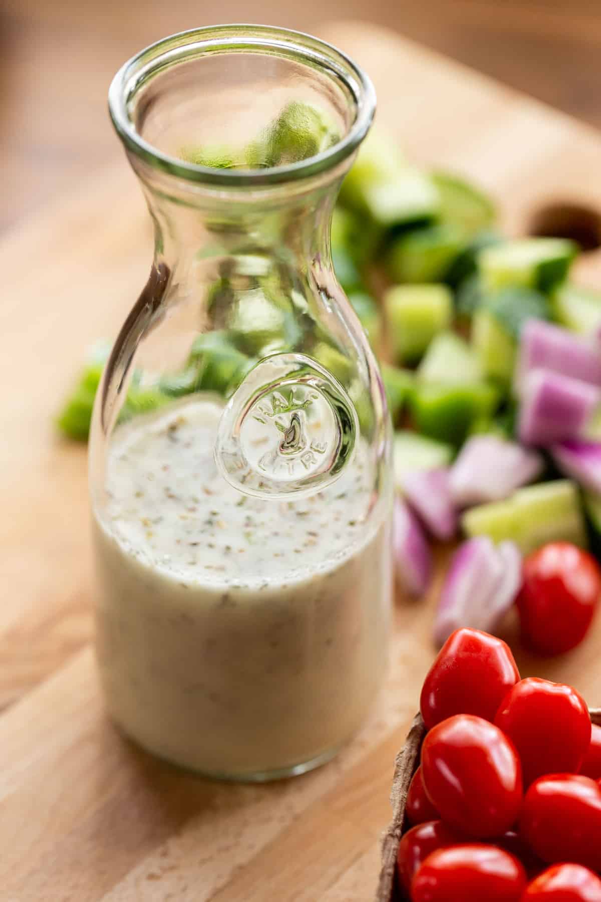 A bottle of Greek salad dressing and fresh vegetables on a wood cutting board.