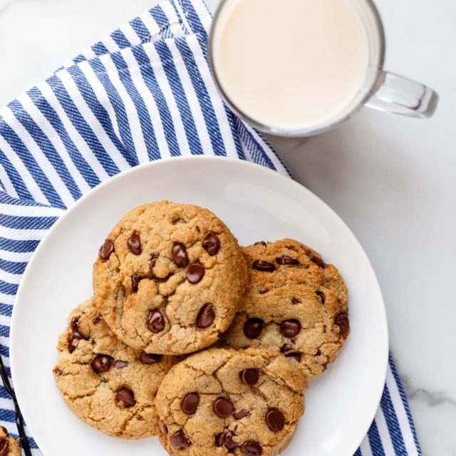Close up of a plate of vegan chocolate chip cookies on a blue striped tea towel with a glass a non-dairy milk on a marble countertop.